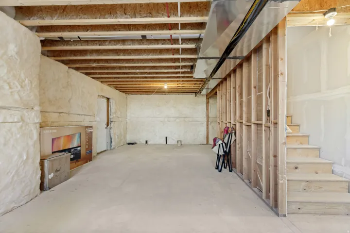 Unfinished basement featuring exposed joists, spray foam insulation, concrete flooring, and an unfinished wooden staircase