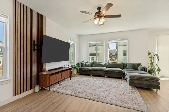 Wood-finish flooring throughout, modern wood slat accent wall, ceiling fan with integrated lighting, multiple windows, and a built-in media console