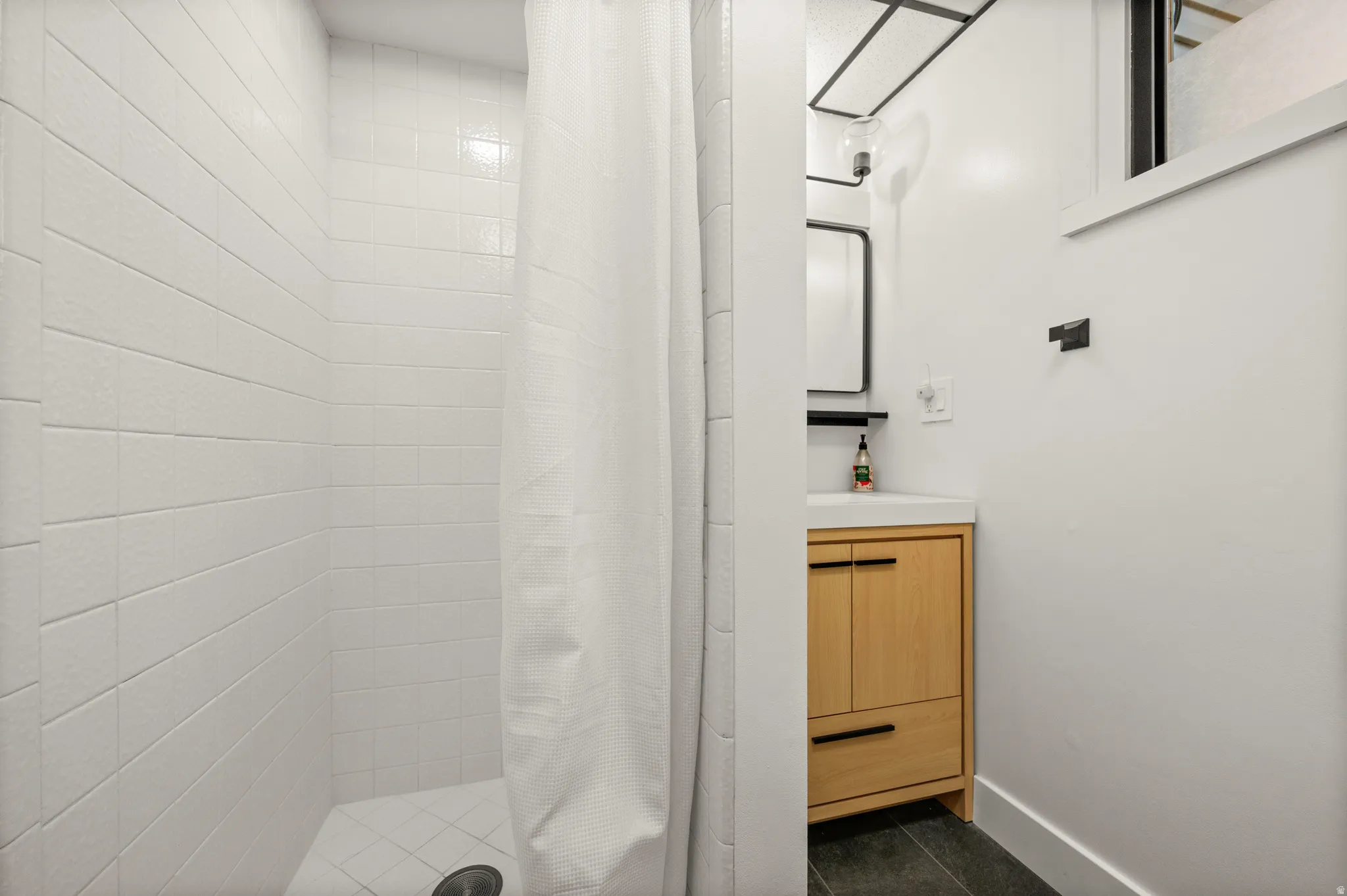 White subway tile shower surround, light wood-finish vanity with integrated sink, black rectangular mirror, matte black hardware, and dark tile flooring