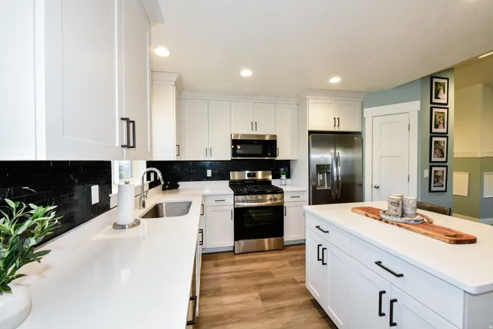 White cabinetry with matte black hardware, stainless steel appliances, black tile backsplash, wood-finish flooring, and a central island with a white countertop