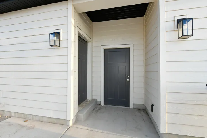 White horizontal siding exterior with black trim, two modern exterior sconces, and a dark paneled entry door
