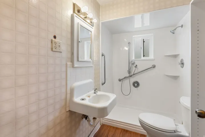 Wall-mounted sink, mirrored medicine cabinet with vanity lighting, walk-in shower with grab bars and built-in shelving, and wood-finish flooring