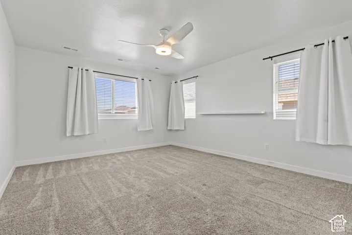 Neutral tone wall paint, textured carpet flooring, contemporary ceiling fan with integrated lighting, multiple windows with blinds, and a built-in wall shelf