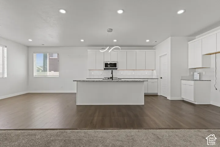Open-concept living area with wood-finish flooring, recessed lighting, and a kitchen featuring white cabinetry, a central island with a sink, and stone countertops