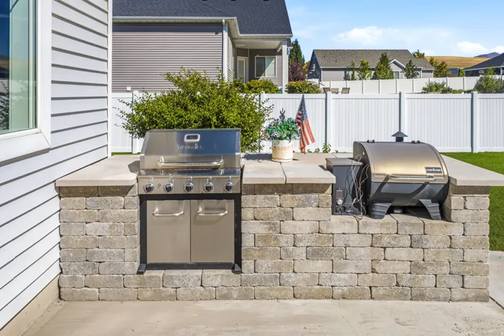 Outdoor kitchen featuring a built-in stainless steel grill, stone block base, and a white privacy fence