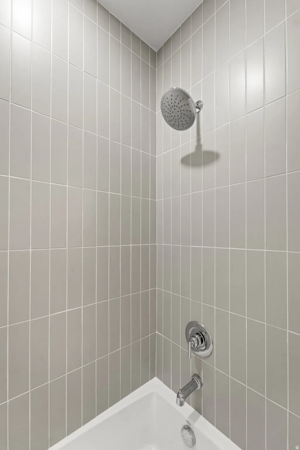 White bathtub with chrome fixtures, a rainfall shower head, and light-colored vertical tile surround