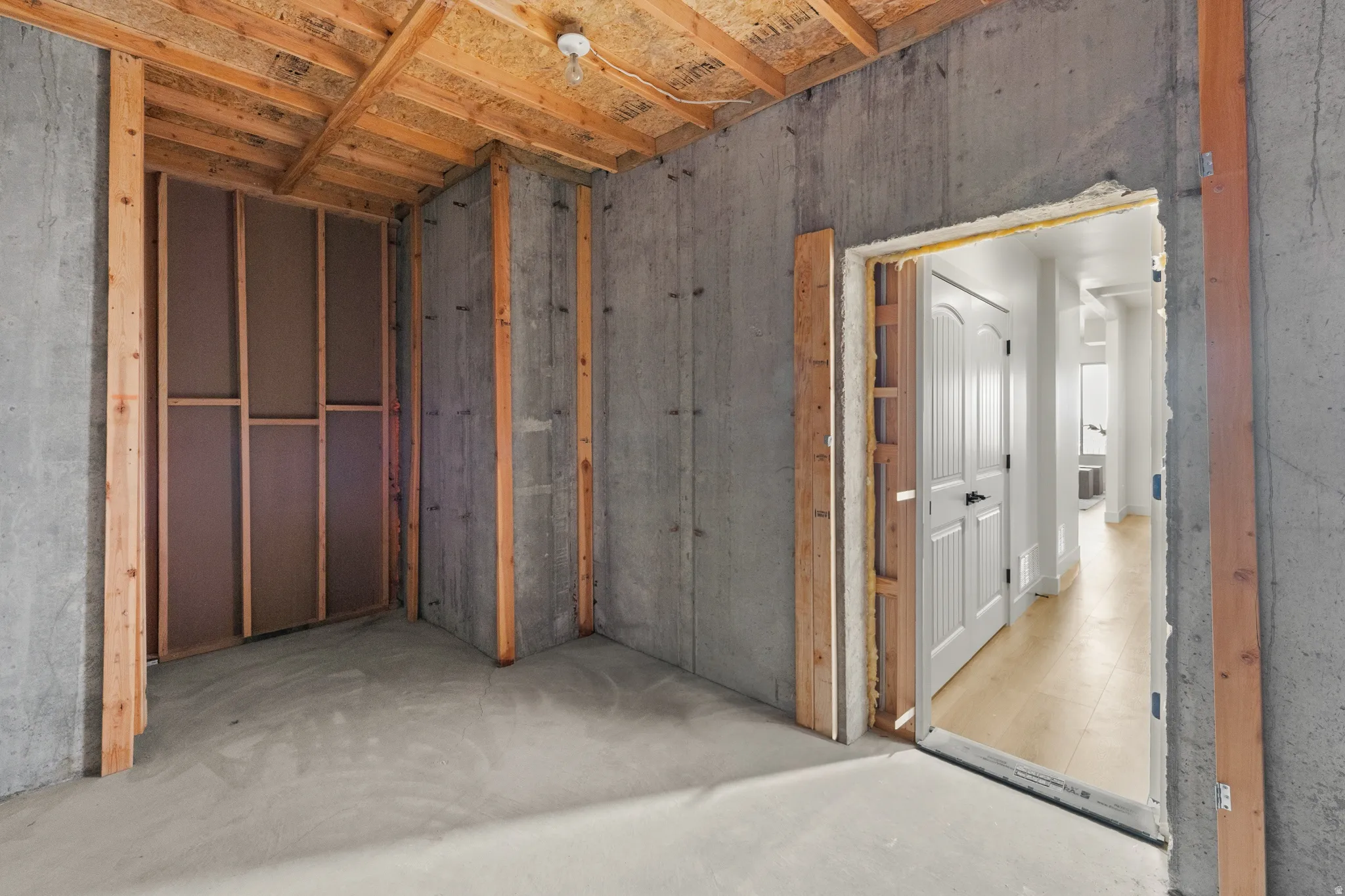 Unfinished concrete walls with exposed wood framing, concrete flooring, and an adjacent finished space with wood-finish flooring and a white paneled door