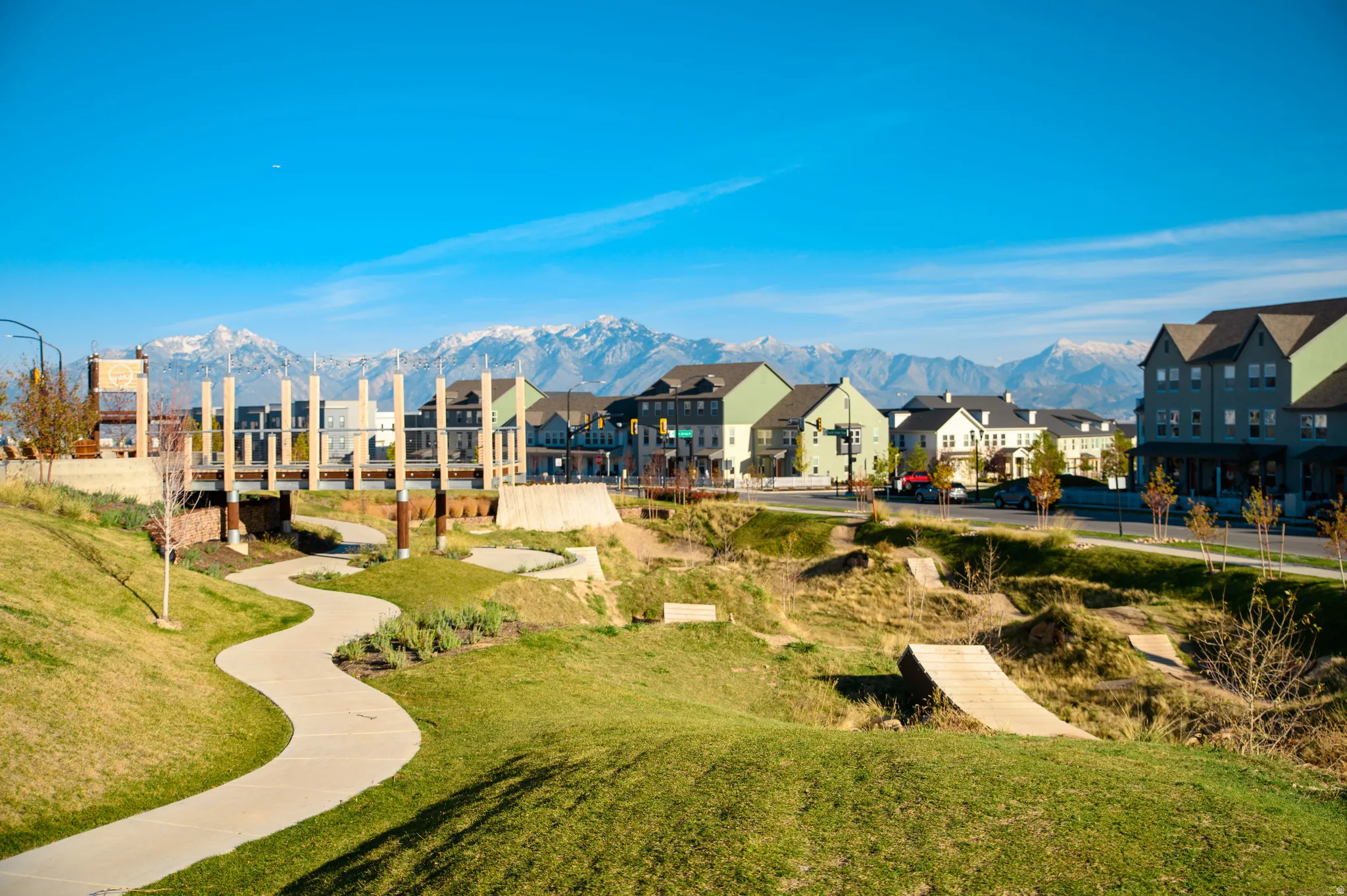 Winding pedestrian path through a landscaped green space with a pedestrian bridge, featuring a residential community and snow-capped mountains in the background