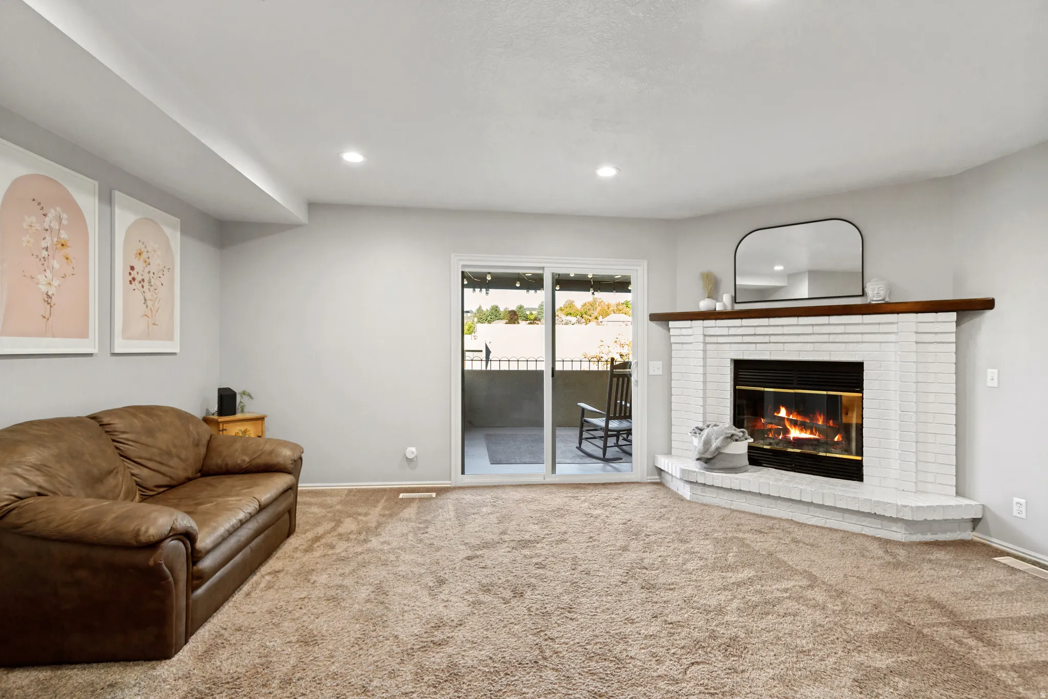Recessed lighting, light-toned carpeting, white brick fireplace with dark wood mantle, and sliding glass door to exterior balcony