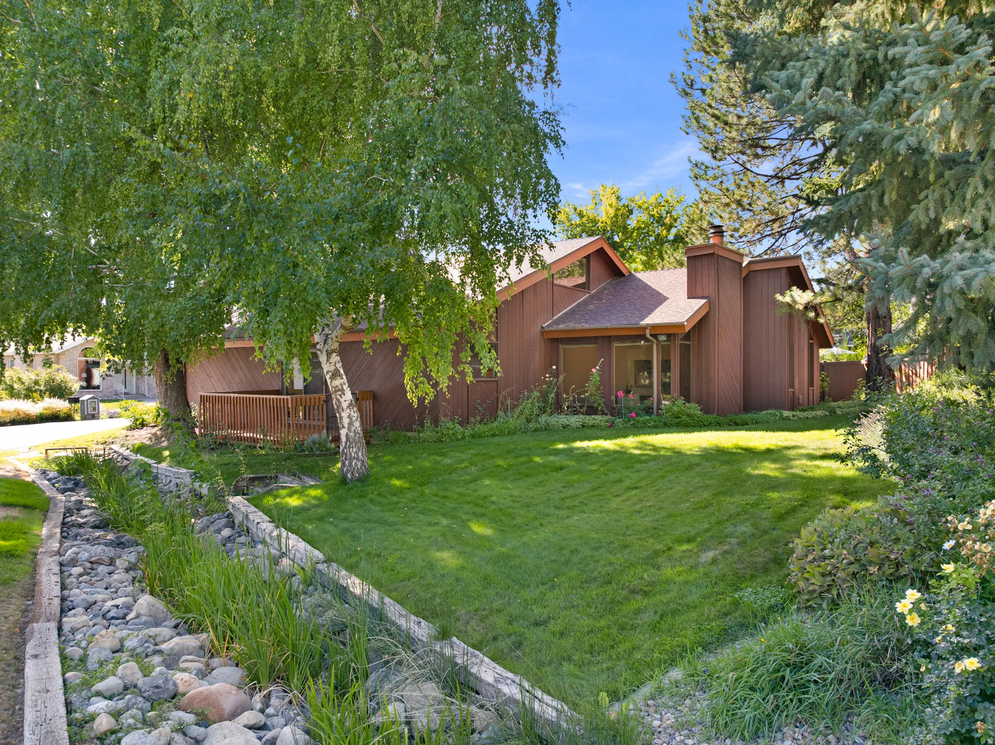 Brown vertical siding, a wood deck, a pitched roof, a stone-lined channel, and mature trees define the exterior