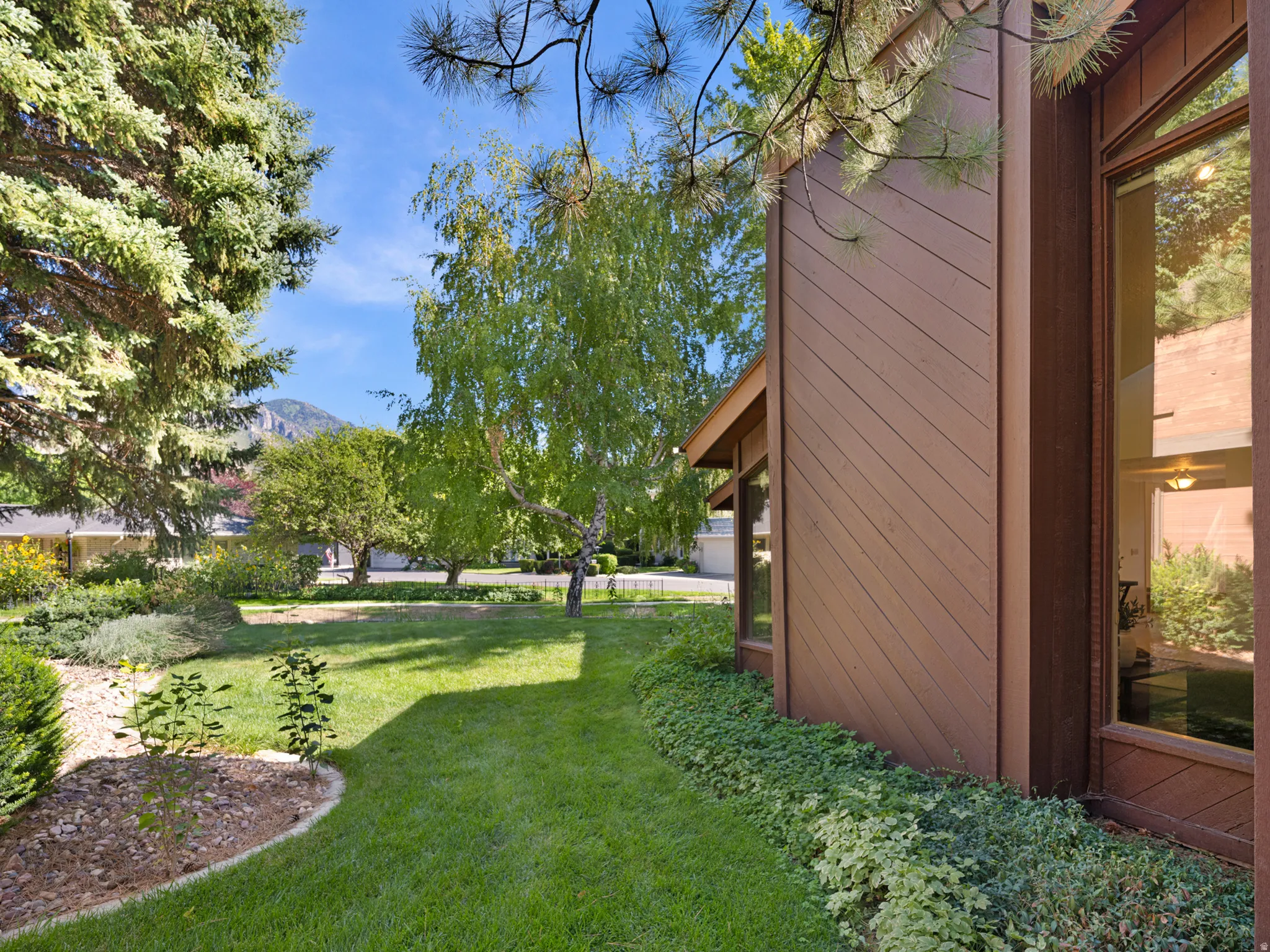 Wood-sided exterior with large window, surrounded by mature trees, manicured lawn, and landscaped garden beds