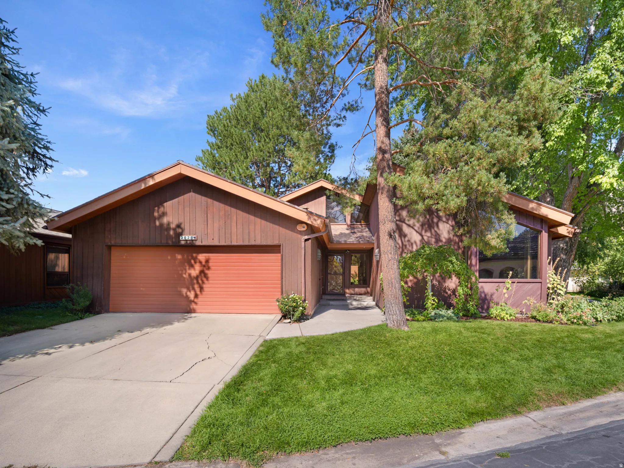 Wood-sided exterior with an attached garage, large picture windows, and a mature tree providing natural shade