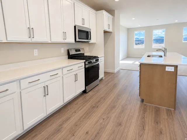 Kitchen featuring white cabinetry, light-toned countertops, stainless steel appliances, a center island with an undermount sink, and wood-finish flooring