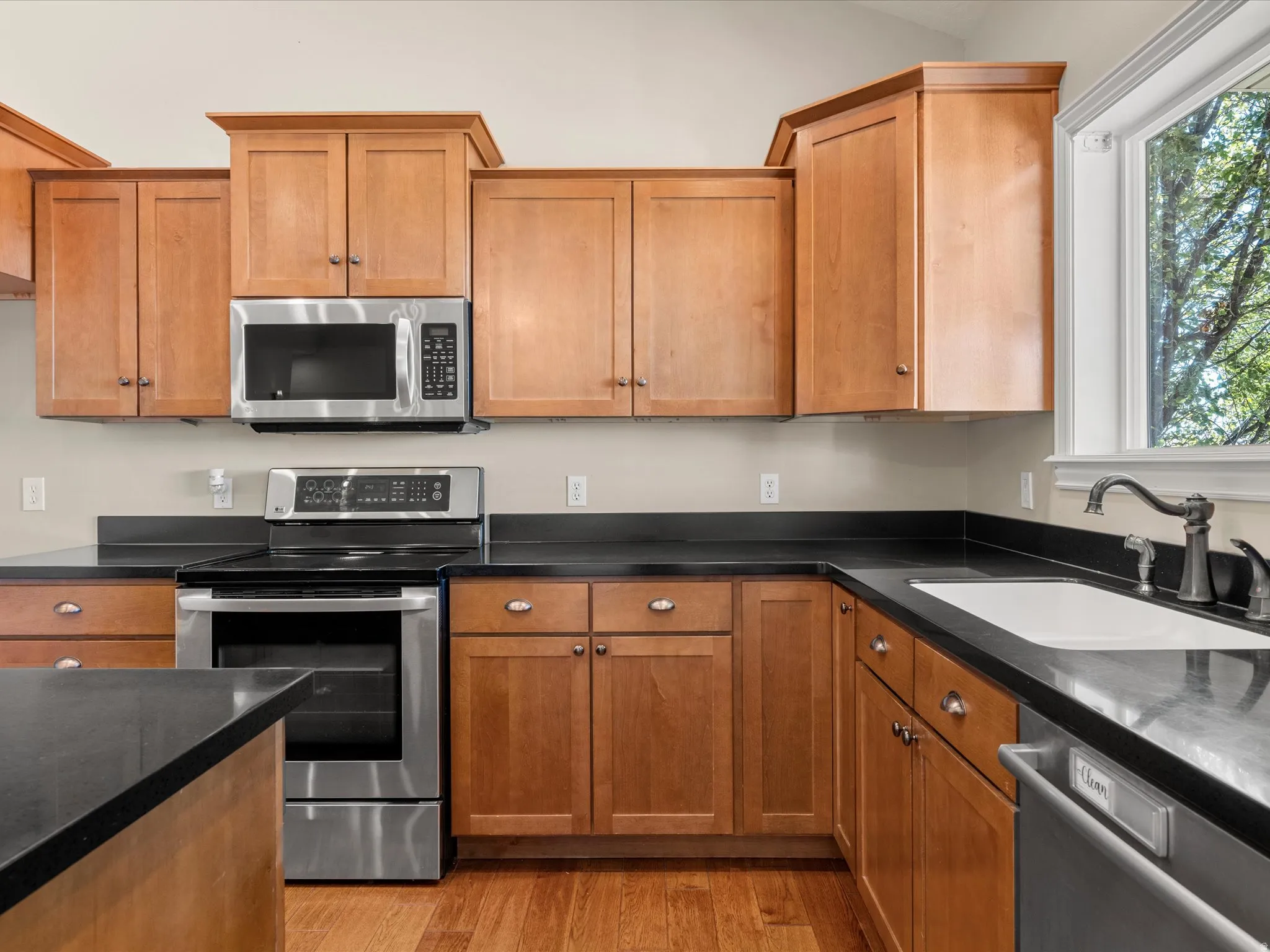 Chef's kitchen featuring warm wood cabinetry with shaker-style doors, stainless steel appliances including a slide-in range and microwave, black quartz countertops, a undermount sink with oil-rubbed bronze faucet, and wood-finish flooring throughout