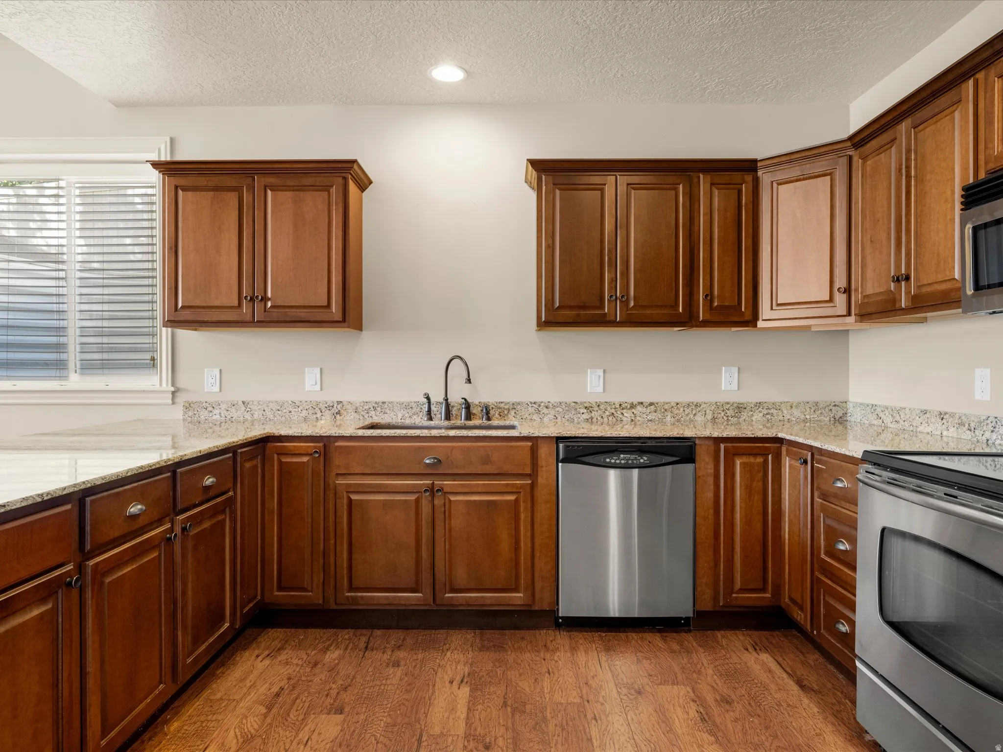 Kitchen featuring wood-finish flooring, L-shaped cabinetry, light-toned countertops, a stainless steel sink with a gooseneck faucet, and stainless steel appliances