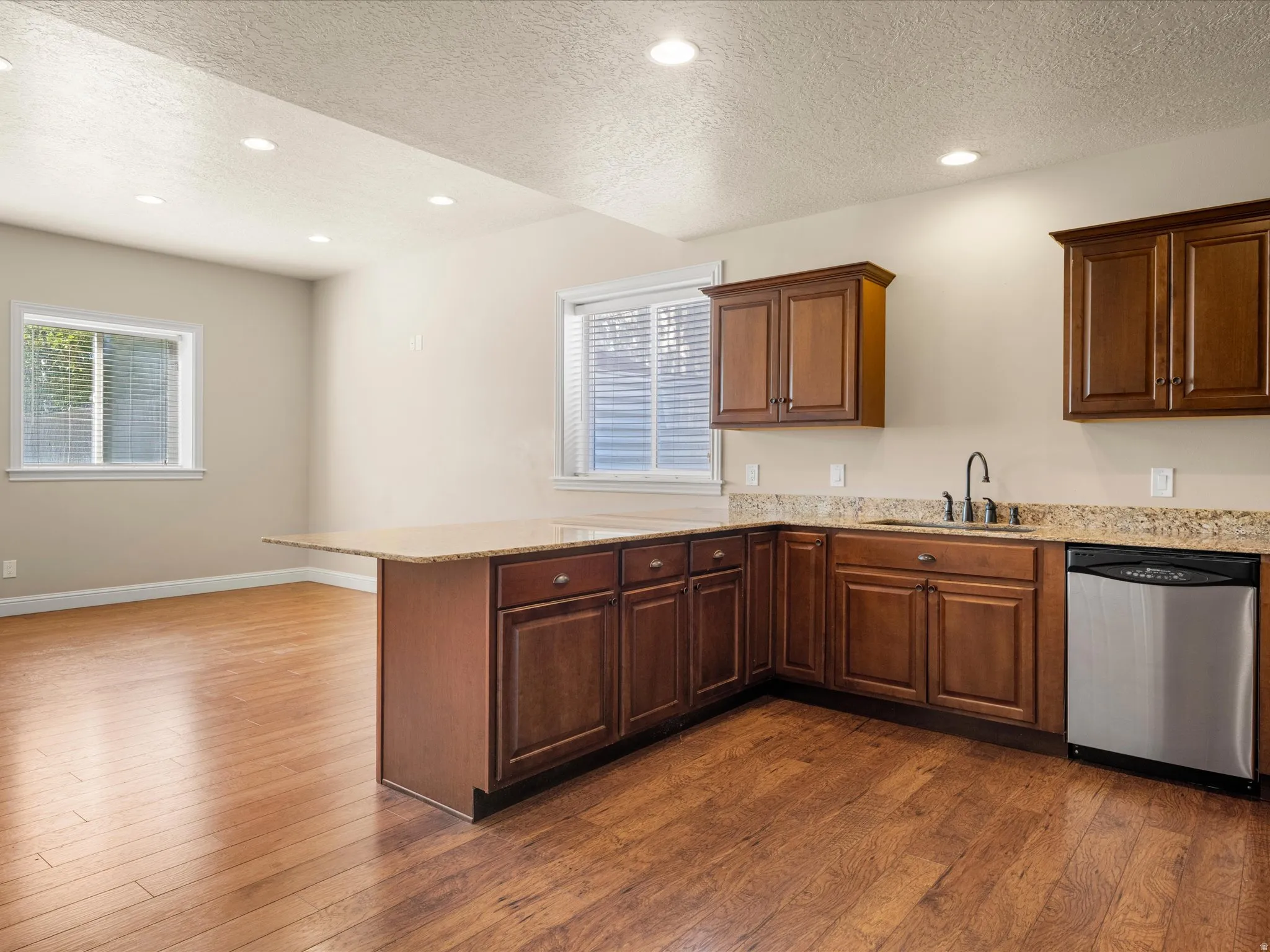 Open-concept kitchen and living area featuring rich wood cabinetry with abundant storage, granite countertops, stainless steel appliances including a dishwasher, recessed lighting throughout, and warm wood-finish flooring that extends into the adjoining l