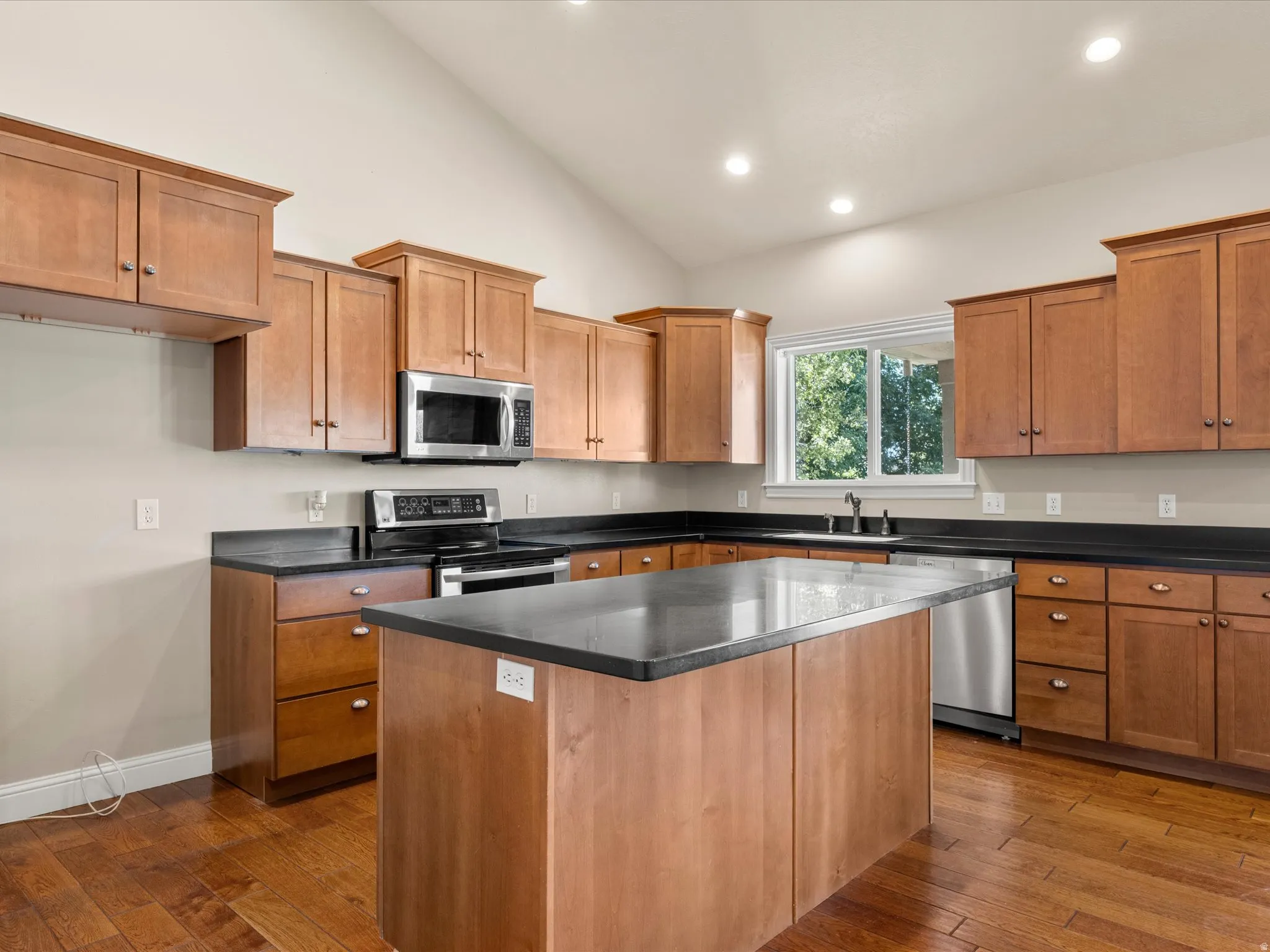 Vaulted ceiling, recessed lighting, wood-finish flooring, wood cabinetry with dark countertops, and stainless steel appliances