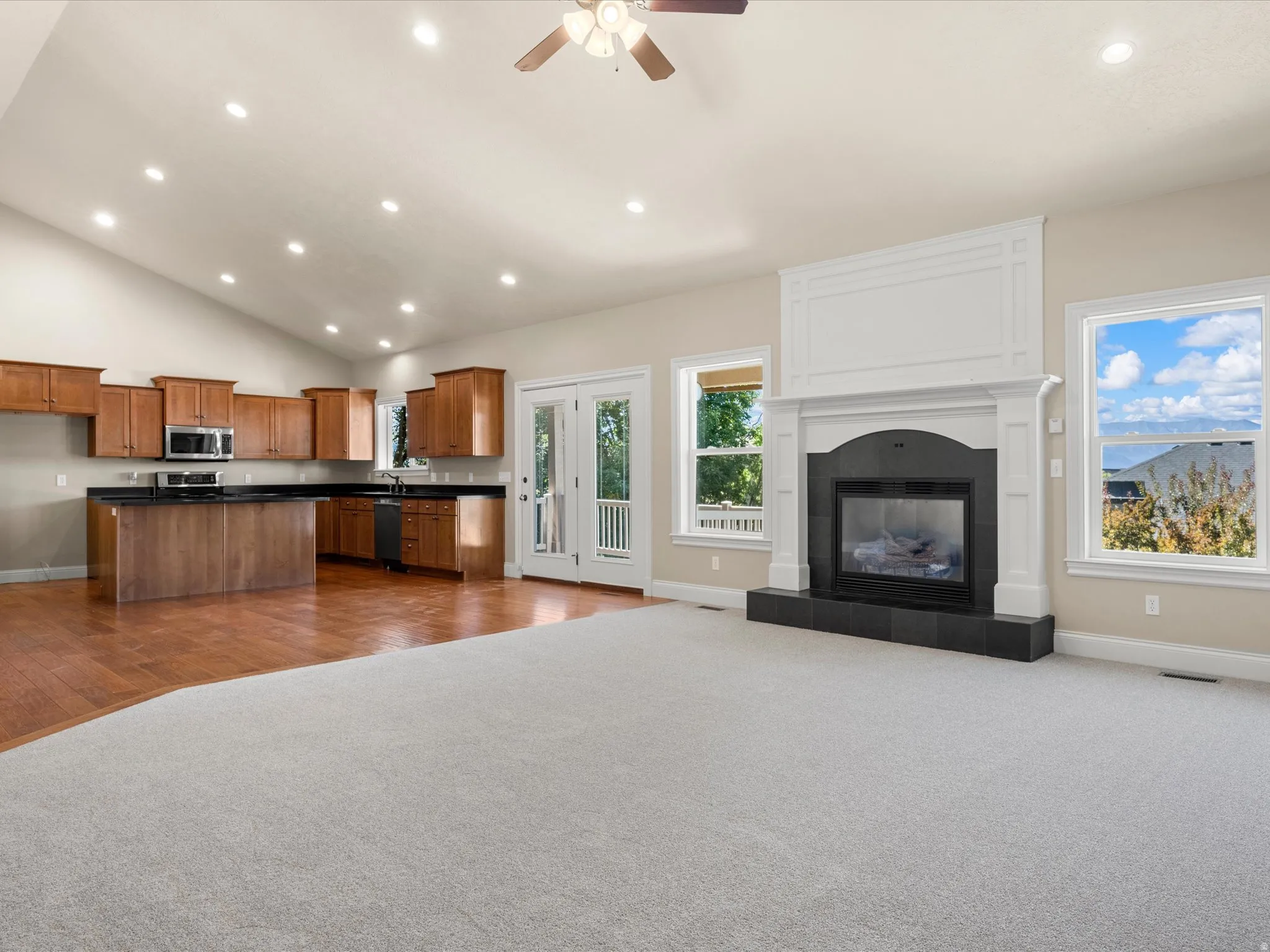 Vaulted ceiling with recessed lighting, wood-finish flooring, white fireplace surround with dark hearth, and wood cabinetry with dark countertops