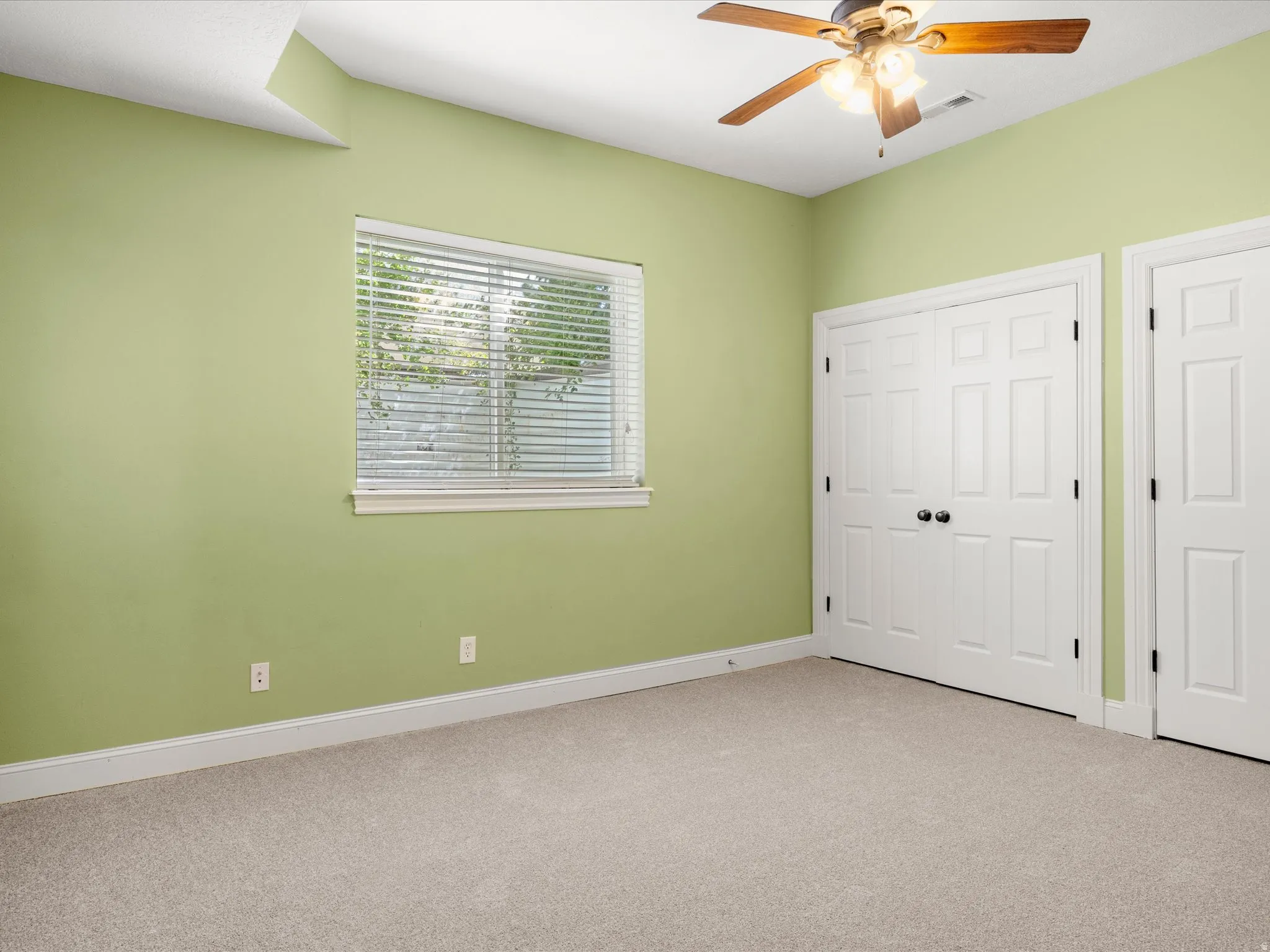 Neutral carpeting, ceiling fan with light fixture, window with blinds, and white bi-fold closet doors