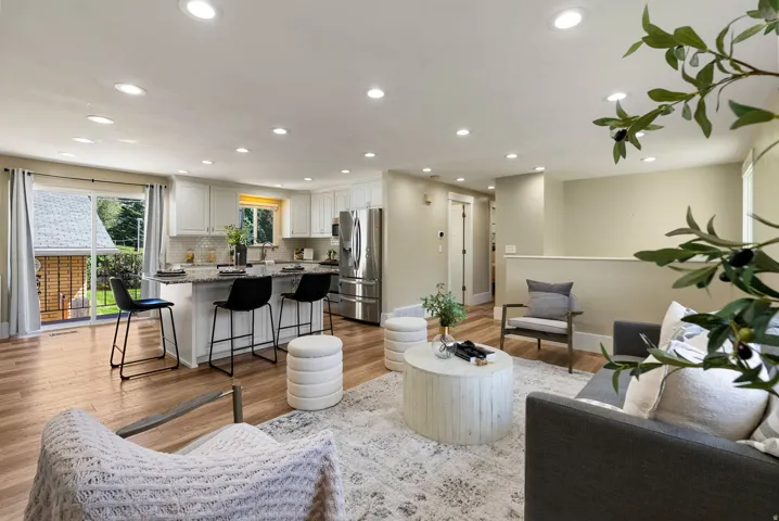 Recessed lighting illuminates the open-concept living space, featuring wood-finish flooring and a kitchen with white cabinetry and stainless steel appliances
