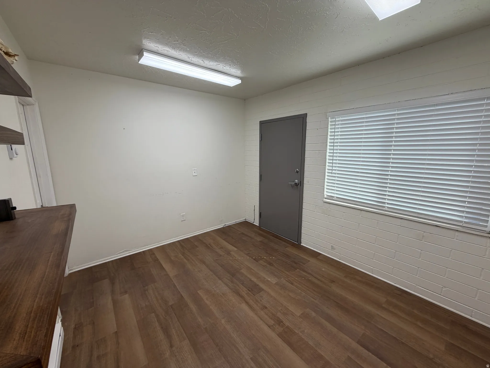 Wood-finish flooring, white painted walls, window with blinds, overhead fluorescent lighting, and a solid gray door