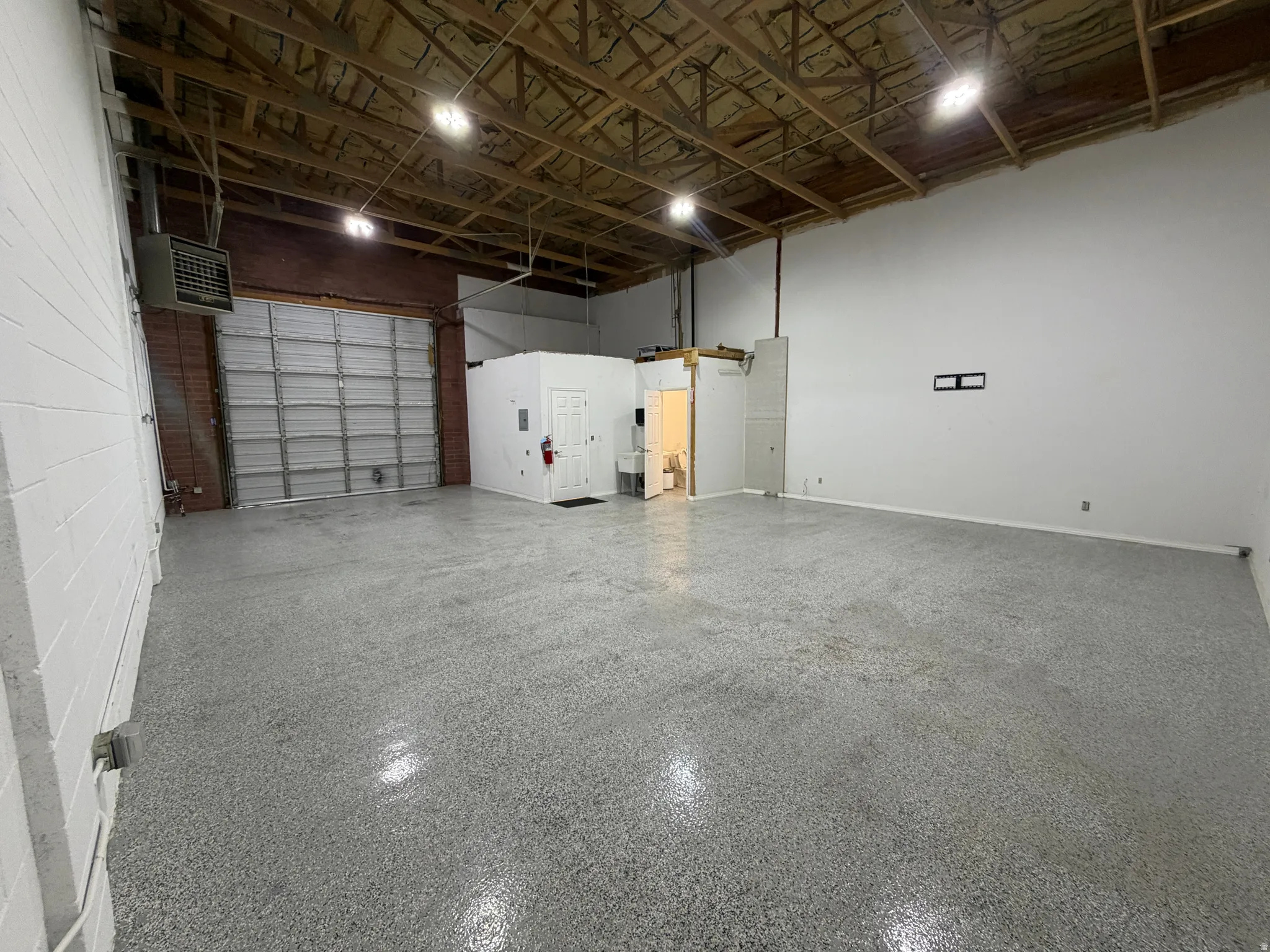 Spacious open area featuring an exposed beam ceiling, concrete flooring, a large roll-up door, white painted brick walls, and recessed lighting