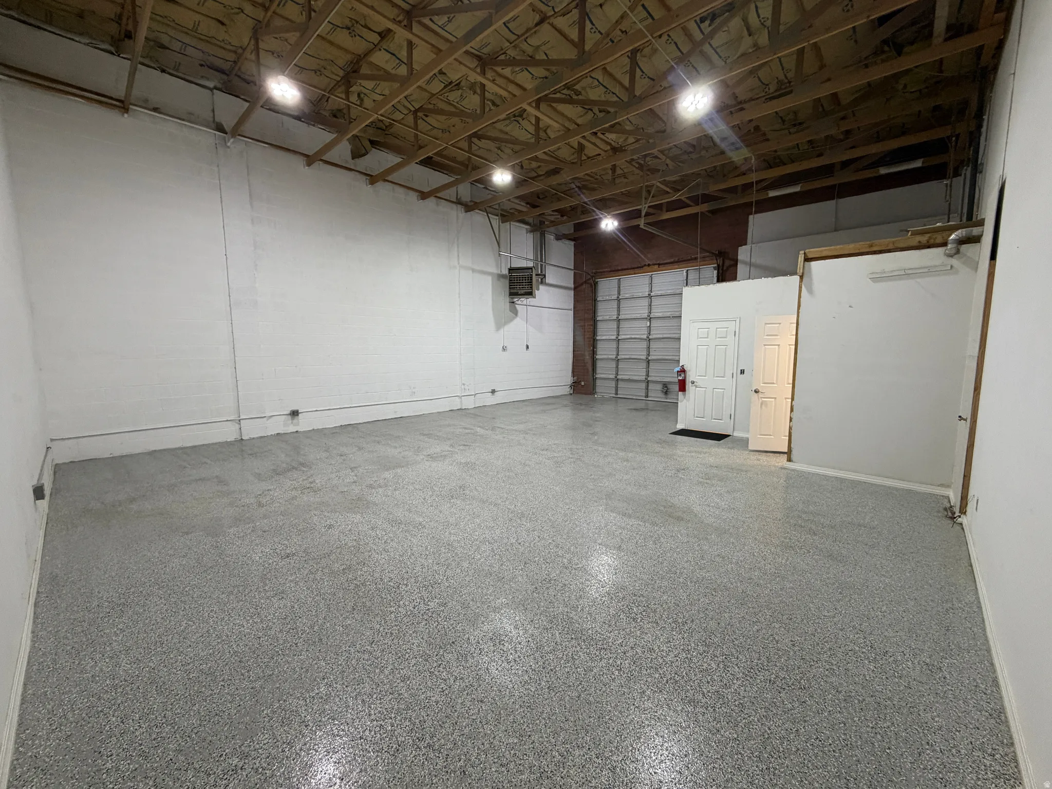 Warehouse space featuring a rolling overhead door, high exposed beam ceiling, white painted walls, and a speckled epoxy floor