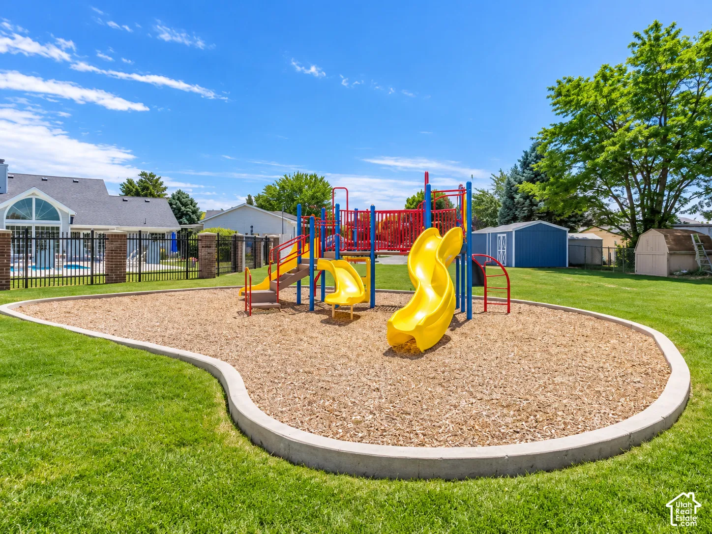 Outdoor play structure with multiple slides and climbing elements, set on a wood chip surface, bordered by a concrete curb and expansive green lawn