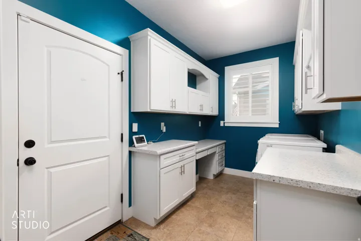 White shaker-style cabinetry with silver hardware, light-toned countertops, built-in desk area, window with white plantation shutters, and textured tile flooring