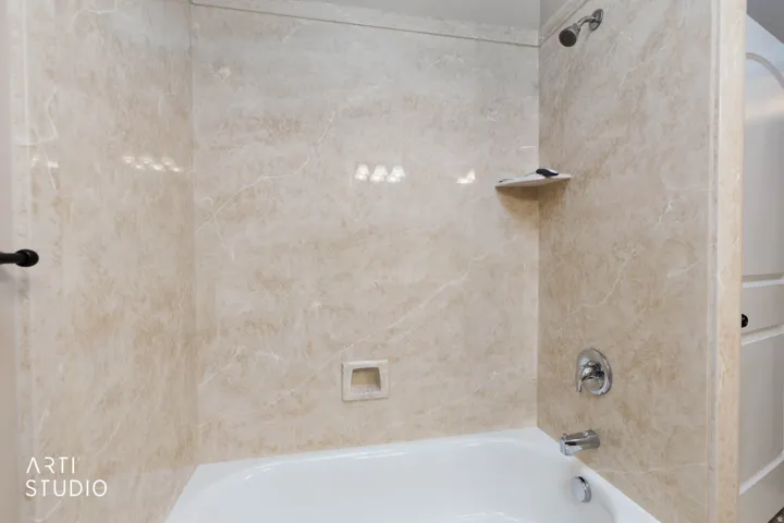 Bathtub with a shower fixture, integrated soap dish, and a corner shelf, all surrounded by a stone-look shower surround
