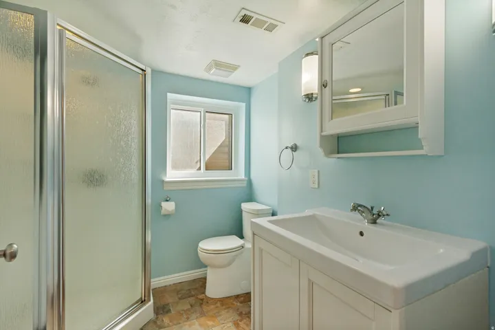 Shower enclosure with frosted glass panels, white vanity with integrated sink, wall-mounted mirrored cabinet, and a window with white trim