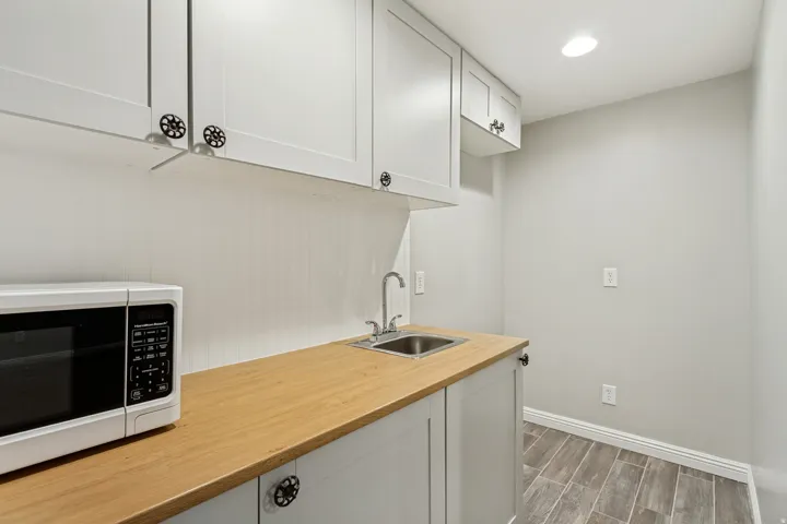 White shaker cabinetry with distinctive hardware, wood-finish countertop, stainless steel sink with chrome faucet, recessed ceiling lighting, and wood-look tile flooring