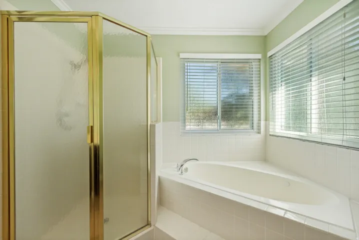 Corner soaking tub with tile surround, dual windows with blinds, and a shower enclosure with a brass-finish frame and frosted glass