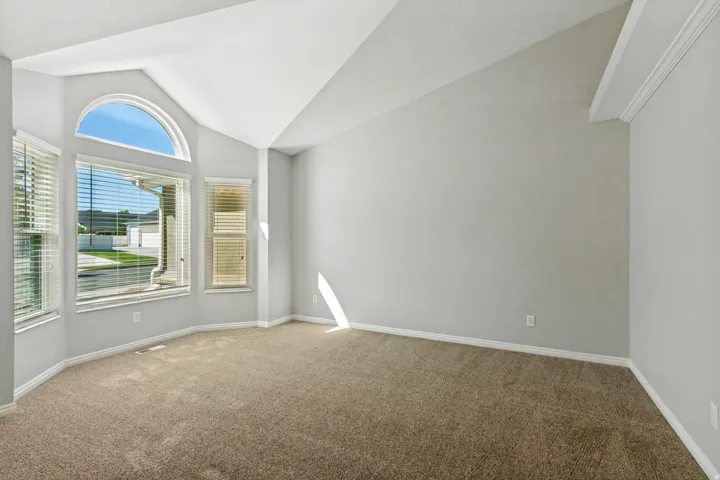 Bright corner bedroom featuring abundant natural light from arched and multi-pane windows, neutral wall finishes, neutral-toned carpet flooring, and clean architectural details with vaulted ceilings