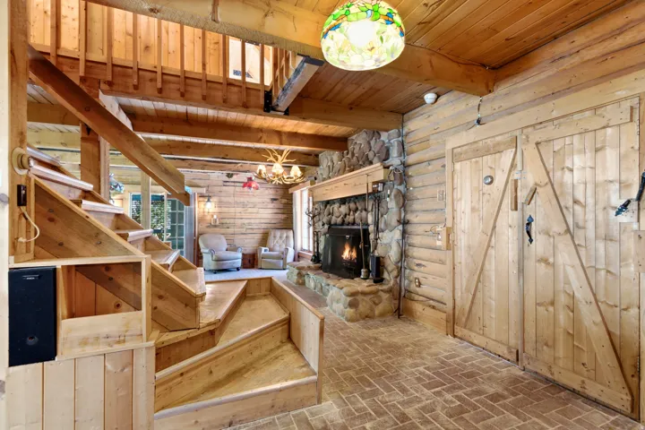 Rustic interior featuring log walls, exposed wood beams, a stone fireplace with a wood mantle, and brick-style flooring