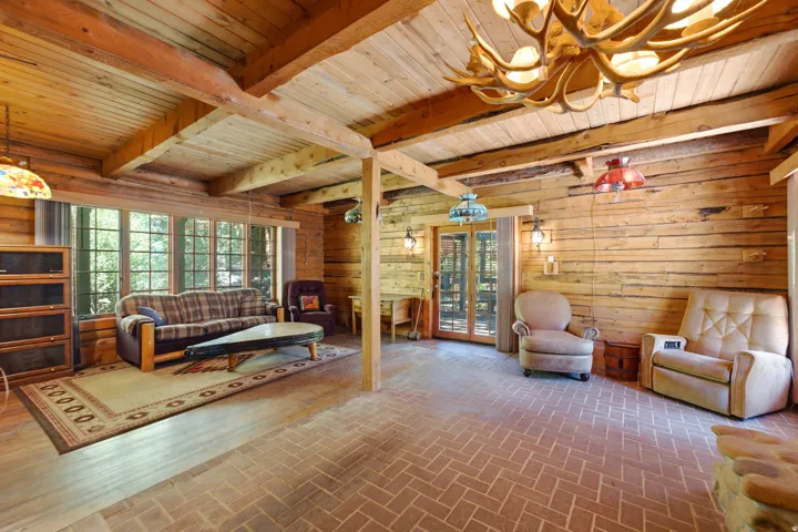 Exposed wooden beam ceiling, wood-paneled walls, expansive windows, brick-patterned flooring, and a decorative antler chandelier