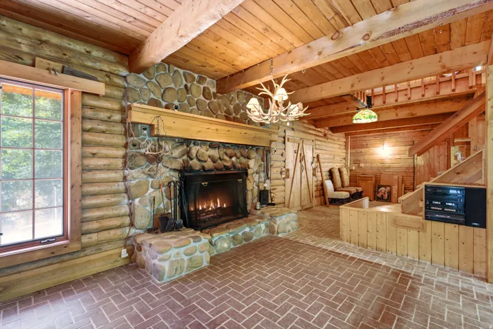 Exposed log walls, large stone fireplace with hearth, wood-finish flooring in a herringbone pattern, exposed wood beam ceiling, and a built-in wooden staircase
