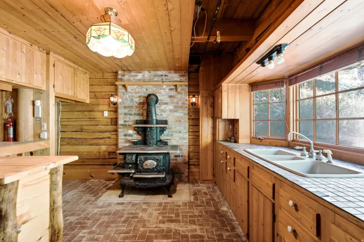 Wood paneling and cabinetry throughout, brick-faced wall with a cast iron stove, tiled countertops, and a dual basin sink beneath a large window