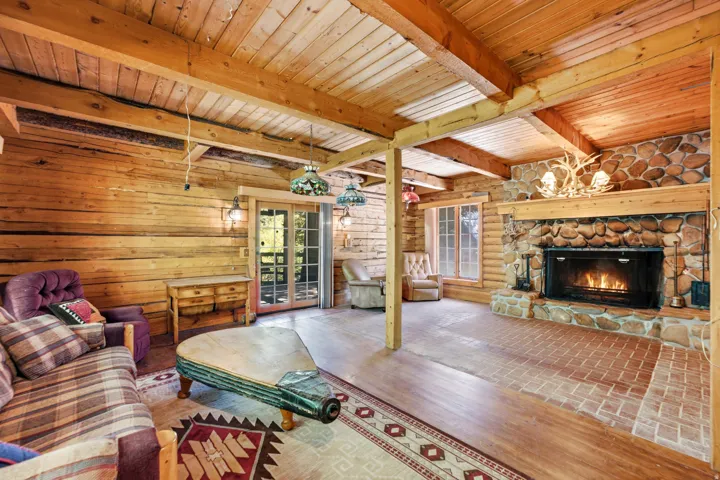 Log walls with exposed wood beams, stone-clad fireplace, brick hearth, wood-finish flooring, and glass-paneled double doors