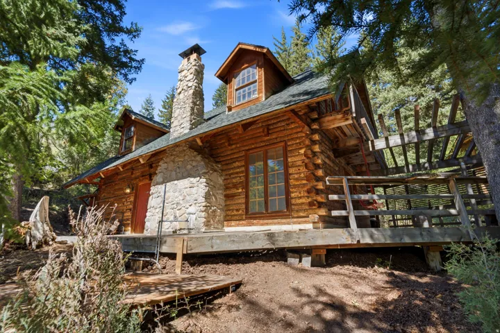 Log cabin exterior featuring a prominent stone chimney, dormer windows, and an elevated wood deck with a pergola