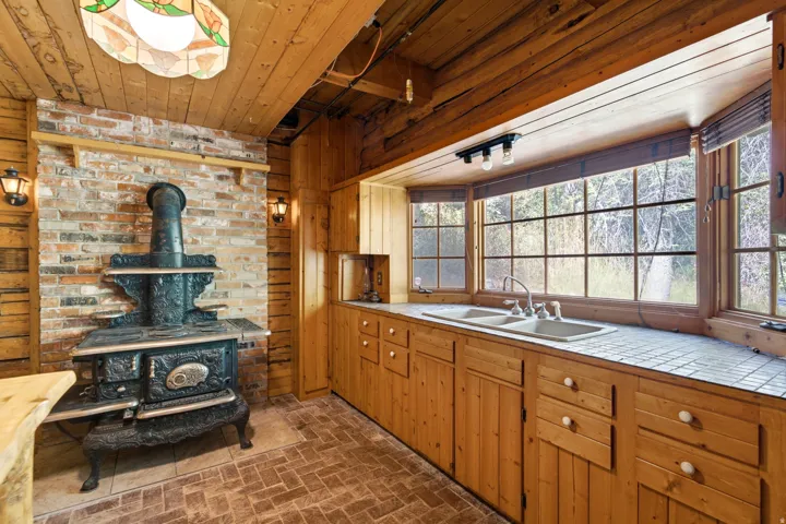 Wood-paneled interior featuring a cast iron stove with a brick surround, a stained glass ceiling fixture, and a bay window with a double basin sink