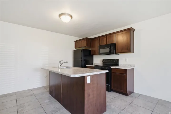 Kitchen featuring dark wood-finish cabinetry, light-toned countertops, black appliances, a central island with an integrated sink, and tile flooring