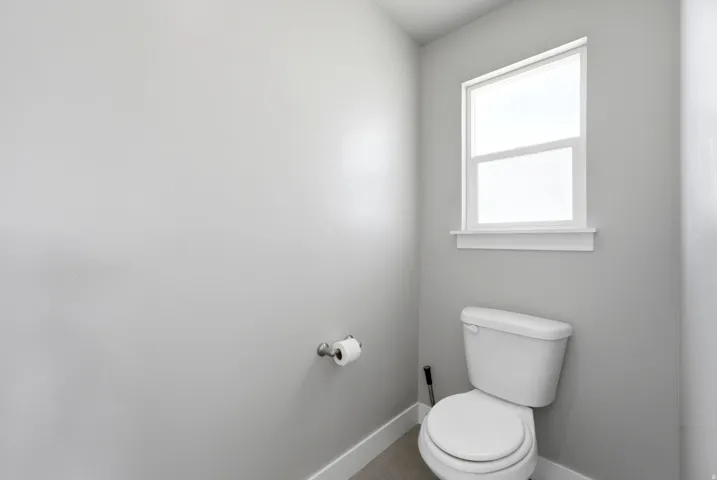 White-framed window, neutral painted walls, toilet with porcelain finish, chrome toilet paper holder, and light-toned flooring