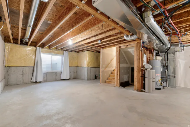 Unfinished basement featuring exposed wood joists, concrete foundation walls, a fixed window, and an exposed staircase