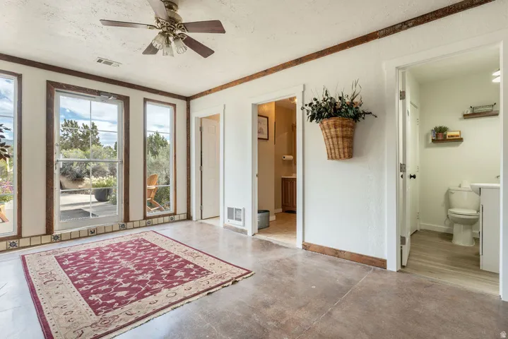 Expansive concrete flooring, ceiling fan, multiple windows with dark wood trim, and direct access to a bathroom with a floating shelf