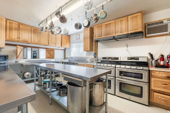 Kitchen featuring multiple stainless steel work surfaces, a pot rack, two double ovens, wood cabinetry, and a window with blinds