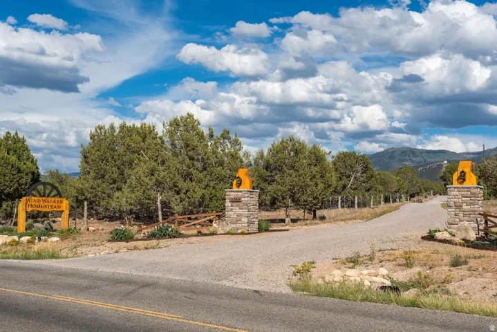 Gravel driveway with stone-clad entry pillars, prominent property signage, natural landscaping, and mountain backdrop