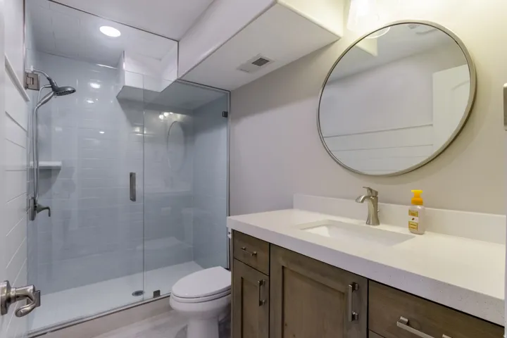 Glass-enclosed shower with subway tile surround, vanity with light-toned wood-finish cabinetry, white countertop, and circular mirror