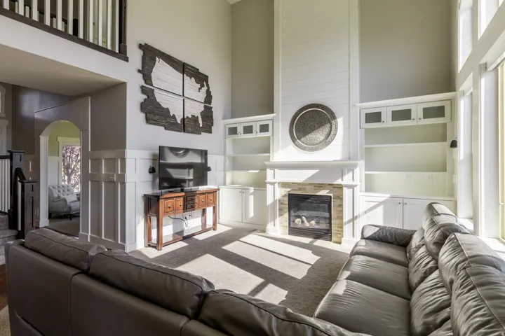 Two-story living area with a stone-surround fireplace, built-in shelving, built-in cabinetry with glass-front uppers, and large windows