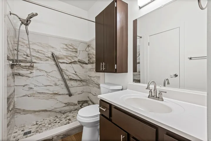 Updated bathroom featuring a walk-in shower with stone-look tile surround and mosaic tile floor, dark wood-finish vanity with integrated sink, brushed nickel faucet, and matching overhead cabinetry.